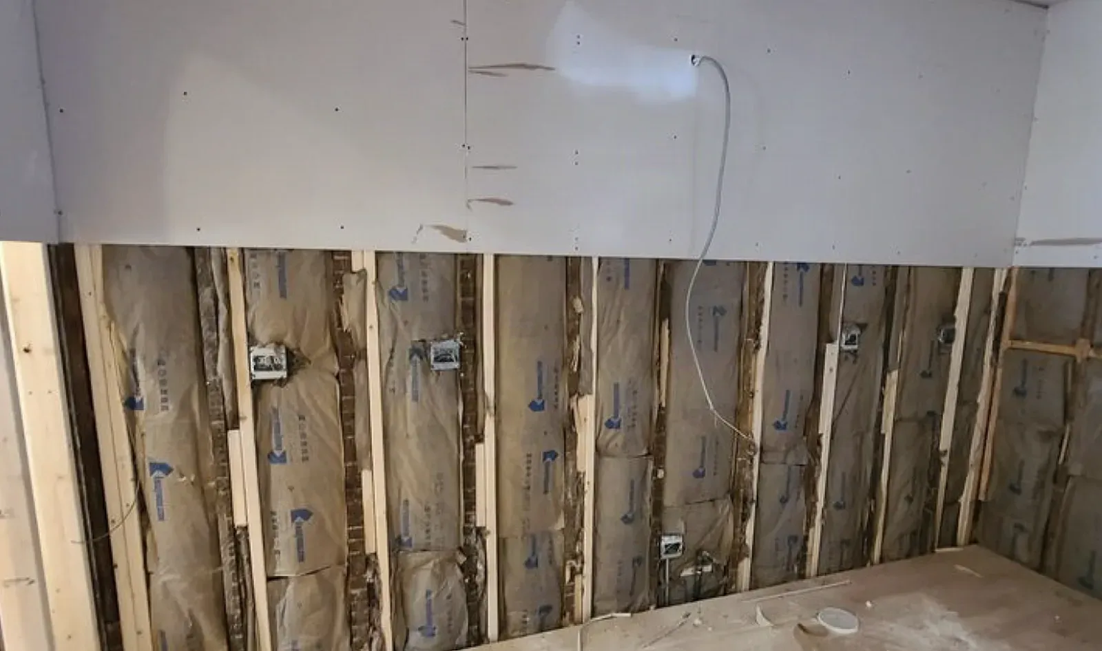Interior wall stripped for demolition, the upper drywall still in place and the lower half removed to expose wood studs, kraft-faced batt insulation, and electrical boxes