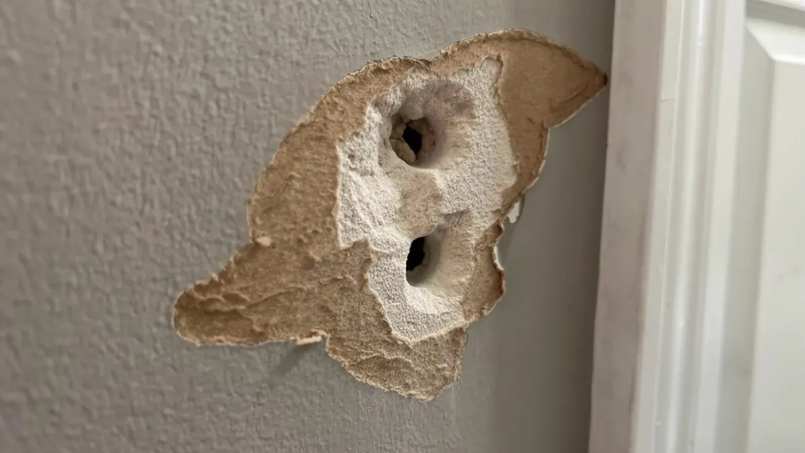Impact hole in an interior drywall wall beside a door casing, torn paper facing peeled back exposing the gypsum core