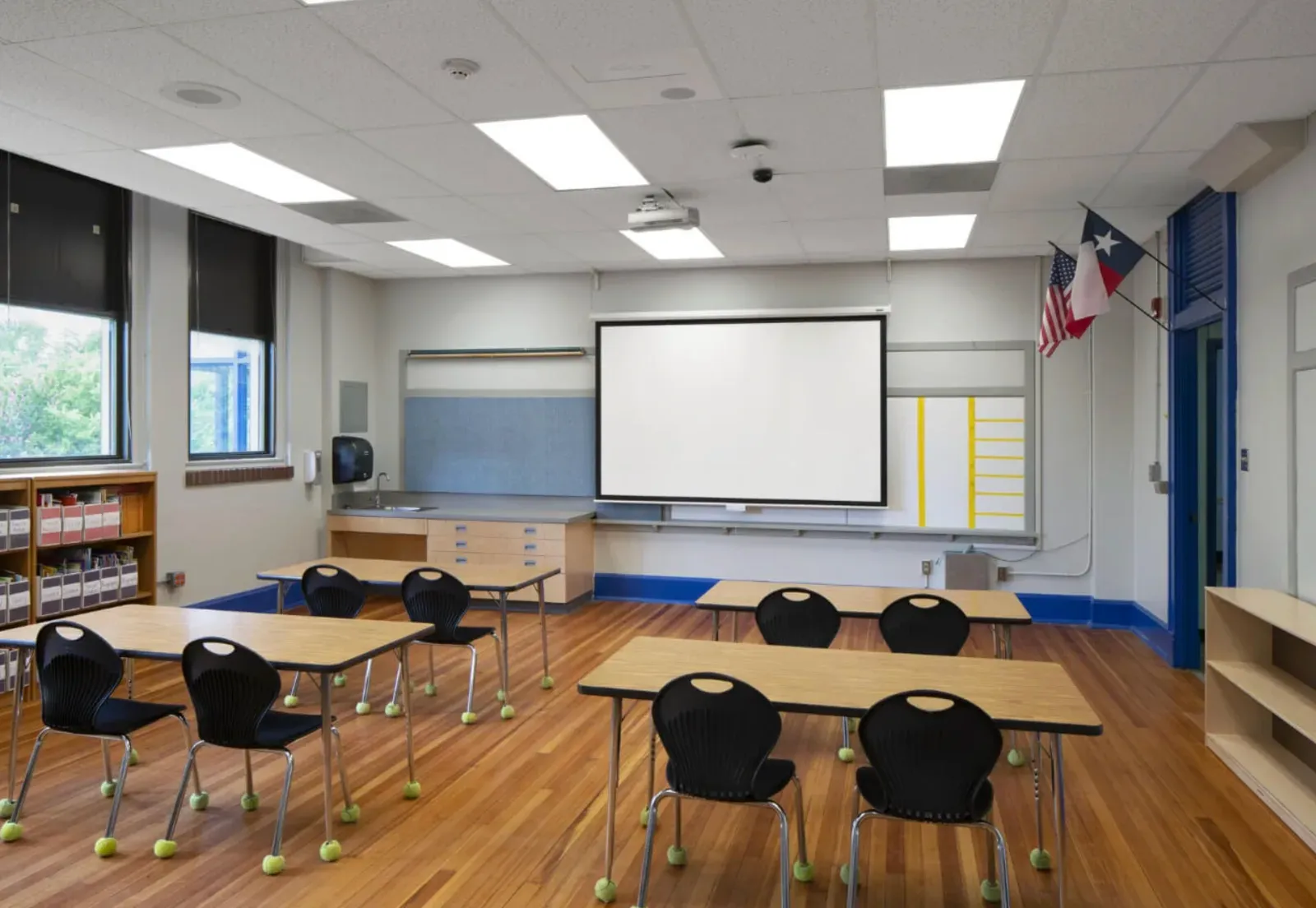 Classroom with finished painted drywall walls, a suspended acoustic-tile ceiling, a pull-down projection screen, wall-mounted casework, and student tables on a wood floor