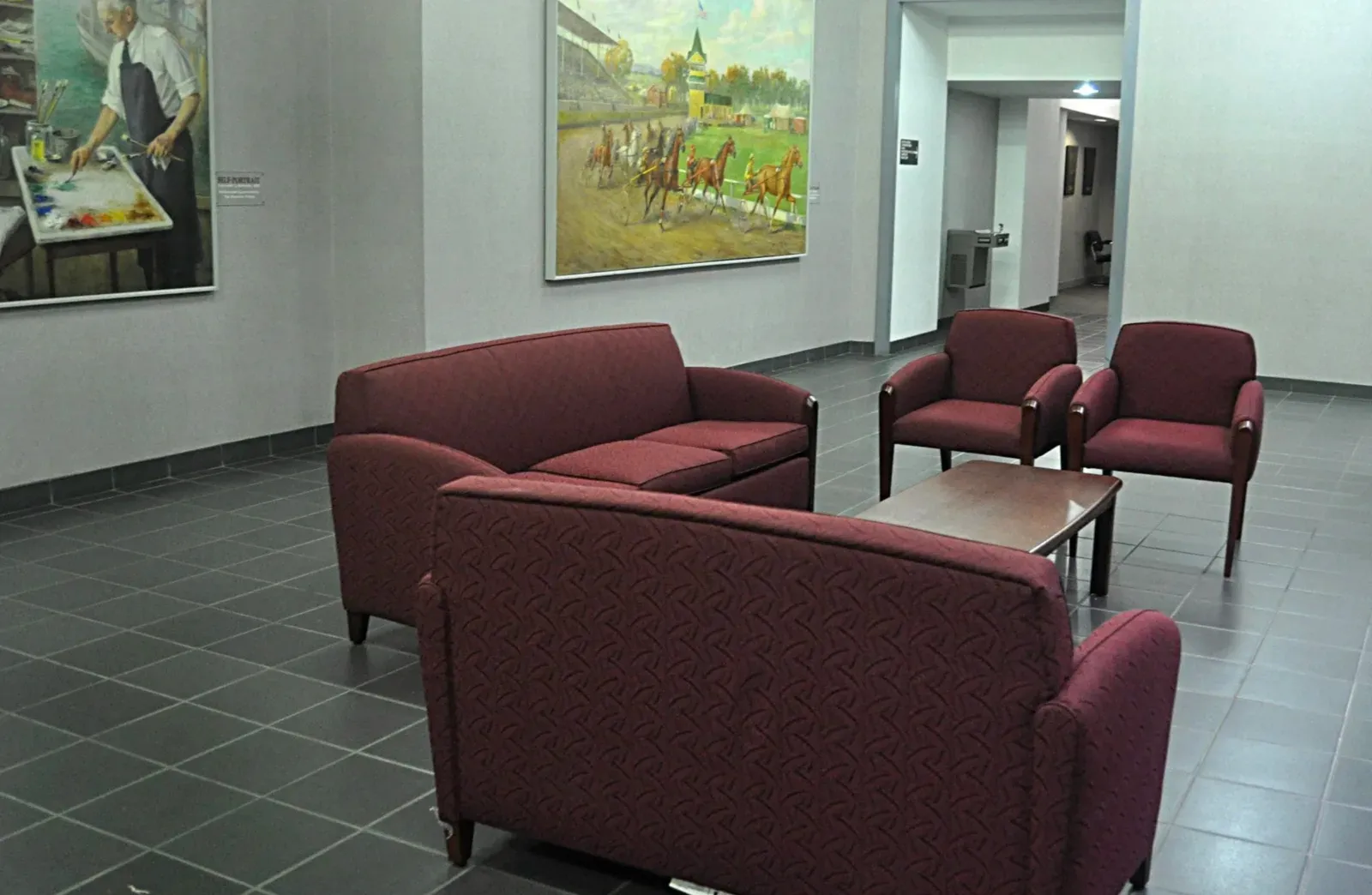 Norwalk City Hall gallery with WPA-era murals hung on smooth plaster-finished walls, a burgundy seating group on gray tile, and a finished corridor beyond