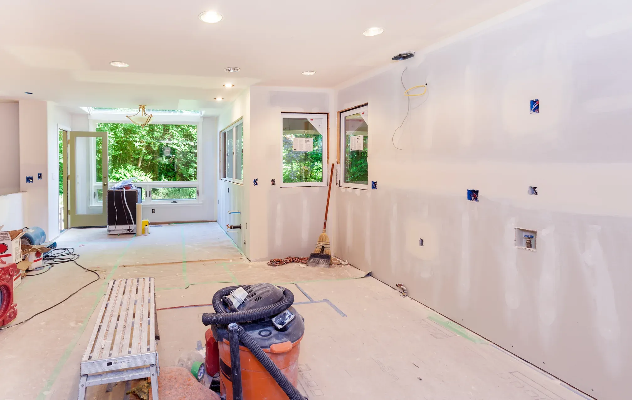Long open interior under construction with freshly hung and taped drywall on the walls and ceiling, recessed can lights, a bank of windows and a glazed exterior door onto green woods, and a shop vacuum and walk board on the bare subfloor